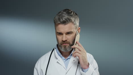 Caucasian-good-looking-grey-haired-male-medic-in-white-gown-speaking-on-the-mobile-phone-on-the-gray-background.-Close-up.